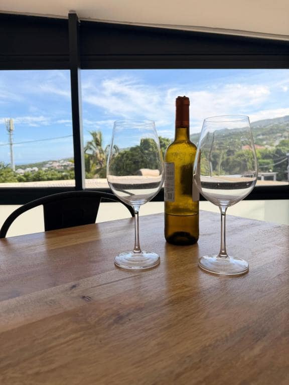 Deux verres à vin sur table bois, vue littoral derrière la baie · © ATÈRLA