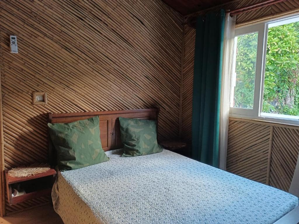 Chambre aux murs de bambou tressé, tête de lit bois et coussins verts · © La baie rose