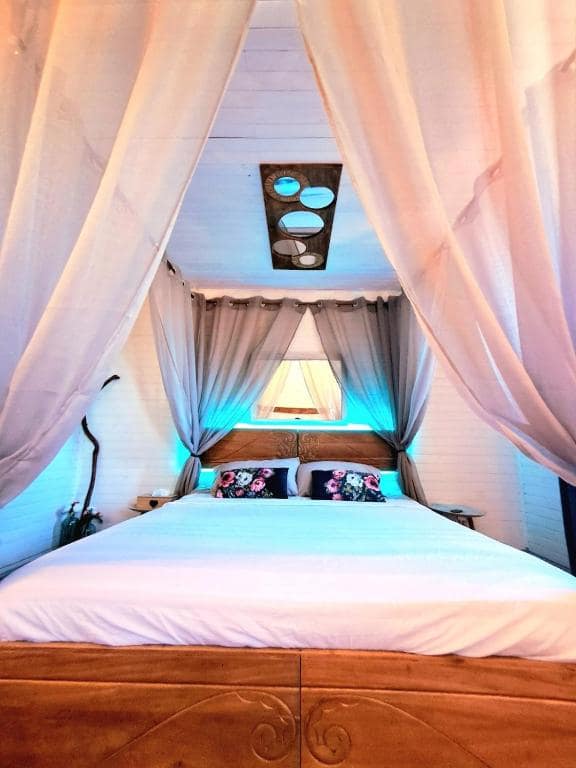 Chambre lit à baldaquin, lambris blanc et coussins fleuris · © l'Antre Temps
