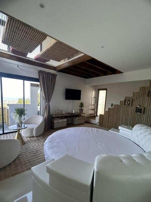 Chambre principale avec grand lit rond blanc, TV et baies sur l'extérieur · © MaNoa
