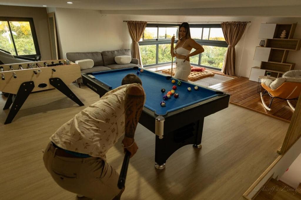 Salle de jeux avec billard bleu, baby-foot latéral et grande baie · © MaNoa