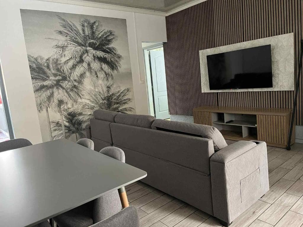 Salon avec canapé gris, télévision et fresque palmiers en noir et blanc · © Le repaire de la mare