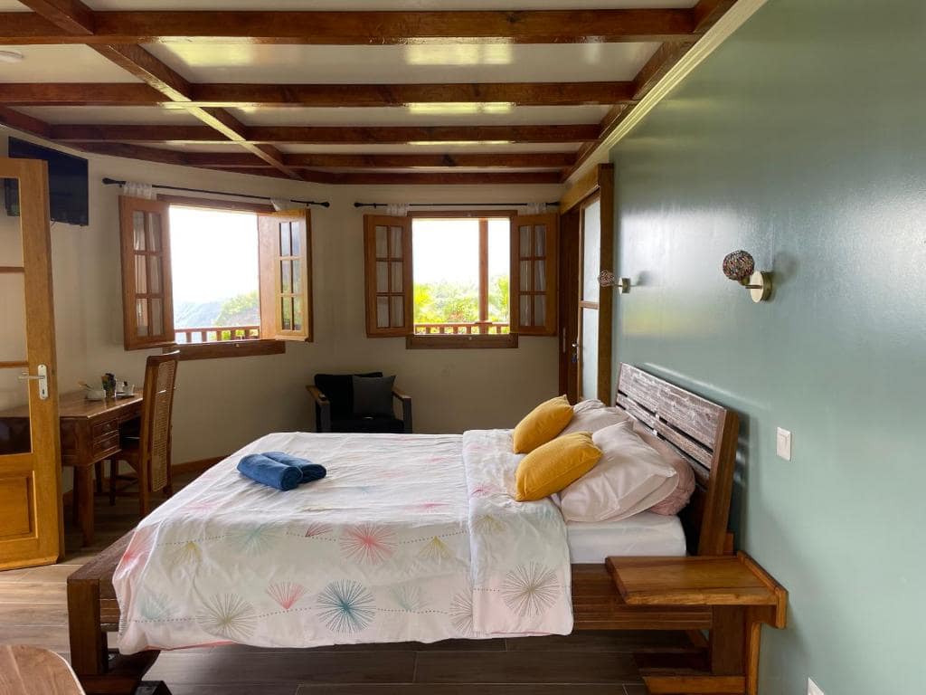 Chambre à grand lit, plafond bois, deux fenêtres à volets ouvertes · © Chambres d'hôtes l'écrin de rose