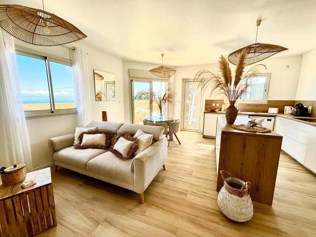 Pièce à vivre : salon beige, cuisine îlot bois, vue mer par la baie · © Ti Cocoon